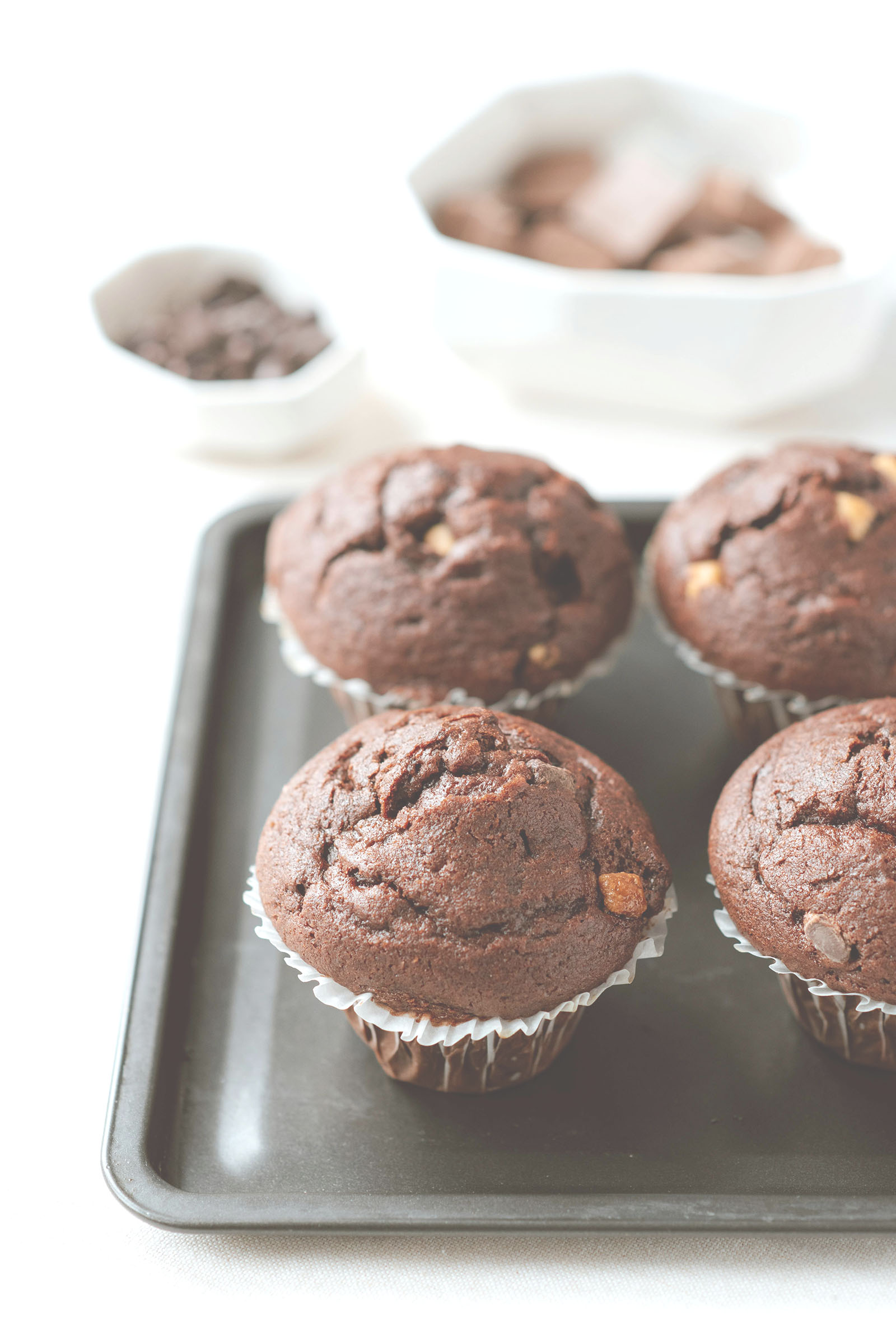 Quatre muffins au chocolat moelleux disposés sur une plaque de cuisson, avec en arrière-plan des morceaux et copeaux de chocolat.